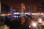 A night-time scene of a dock containing a number of moored canal-boats to left and right and railings around the edges. At the front is part of a lock gate and steps leading down to the water.  Around most of the dock are multi-storey modern buildings, some with lighted ground-floors and seats and decorative objects outside.  The most prominent of these, at the far end, is a twenty-storey building with curving facades.