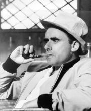 A black and white photo of Henri-Georges Clouzot taken from a side. Clouzot is depicted sitting downsmoking a pipe and wearing a matching white and hat and suit.