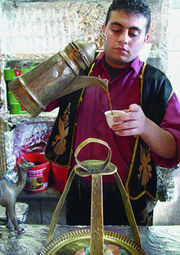 A man with short curly hair, wearing a gold-fringed black vest over a magenta shirt, pours coffee from an ornate, angular container into a small white cup.