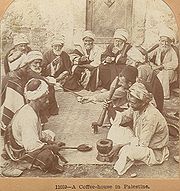 Monochrome photograph of men with turbans and facial hair reclining on a mat. In the foreground a man uses a mortar and pestle, while the men in the back have cups in their hands.