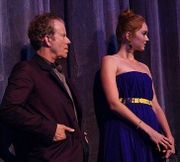 A red-haired woman in a long blue dress standing next to a shorter, Caucasian man with salt and pepper hair, wearing a dark suit