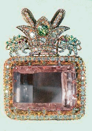 A large rectangular pink multifaceted gemstone, set in a decorative surround. The decoration includes a row of small clear faceted gemstones around the main gem's perimeter, and clusters of gems forming a crest on one side. The crest comprises a three-pointed crown faced by two unidentifiable animals.