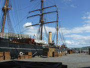  Partial view of a ship moored to a quayside. Prominent visible features are a mast with three crossbeams, two smaller masts, a funnel, a lifeboat and rigging. Packing cases are lined up on the quay, and a gangplank with "RRS Discovery" on it leads to the ship.