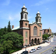 A brick church with two tall, symmetric steeples is seen in front of a city street, to the right of a wooded park.