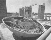 A black and white photo of a round, unfinished building with exposed joists meant to support a future roof; it is surrounded by four towers made of marble and glass.