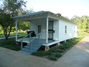 Present-day photograph of a whitewashed house, about 15 feet wide. Four bannistered steps in the foreground lead up to a roofed porch that holds a swing wide enough for two. The front of the house has a door and a single paned window. The visible side of the house, about 30 feet long, has two paned windows.