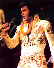 Presley, mutton-chopped and fuller-faced, sings into a handheld microphone. A golden lei is draped around his neck, and he wears a high-collared white jumpsuit resplendent with red, blue, and gold bangles.