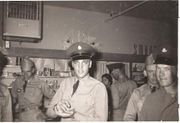 Presley stands in a ship's canteen, wearing a stiff black-brimmed military cap, khaki shirt, and dark tie. His hands are cupped at chest height, holding something. Other soldiers stand to either side of and behind him.