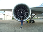  Aircraft engine, forward facing view with a Boeing engineer in front to demonstrate the engine's size.  The engine's large circular intake contains a center hub with a swirl mark, surrounded by multiple curved fan blades.