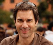 A caucasian male with dark hair, wearing sunglasses on the top of his head, with a brown suede jacket, smiles