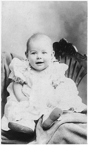  an infant dressed in light colored clothing propped on a chair faces the camera