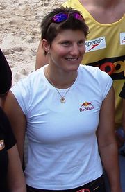 Young smiling woman with short brown hair, wearing a white t-shirt with the Red Bull logo and sleeves pulled back. She also wears a pair of sunglasses on top of her head and a golden chain with a circular pendant around her neck. She is on a beach among a group of people.