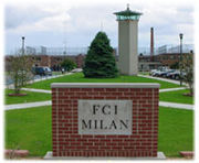 A red brick sign in the foreground says "FCI MILAN". In the back, a solitary green tree is visible atop a grassy mound, with a tower to its right.