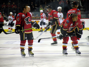 A player in full uniform but without a helmet stands to the right of a teammate who is crouched over.  Both players are looking to their left as several others skate in the background.