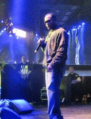 A black man with short black hair. He wears dark sunglasses of an unrecognizable color, a brown jacket, and blue jeans. Two men with brown hair stand far behind him, wearing black shirts with unrecognizable yellow text.