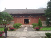 A Chinese temple building that is red. In front of it is a pathway and trees and flowers.