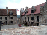 A photograph of two stone buildings with red roofs, surrounding a stone-paved central area. The buildings have entrances on two levels, with wooden stairs outside leading to the doorways on the upper level. An American flag is visible waving in the gap between the buildings.