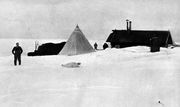  On the right is a long low hut with two chimneys, surrounded by snow drifts. In the centre is a pointed tent and, to the left, a covered mound of stores. Several men are visible, as is a seal.