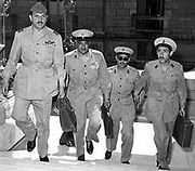 Four men in military uniform, wearing buttoned jackets, wide-rimmed hats, black shoes, and carrying briefcases. They are ascending the stairs of a building.