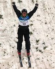 A man skis down a snow-covered slope with his arms in the air and fists closed. He is wearing black equipment with a blue-and-white vest bearing the Olympic rings and the number ten; a black helmet, black-and-white gloves, protective glasses, and red boots attached to black skis.