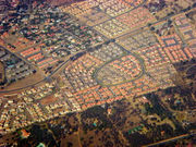 Bird's-eye view of residential neighborhoods. The houses at the bottom third of the picture have much larger yards and more trees. The neighborhood in the center of the picture has a very defined road shaped like a backwards 6, and the houses have orange and grey roofs and are very close to each other. The neighborhood at the top right has multicolor roofs. There is a road cutting through the middle of the picture.