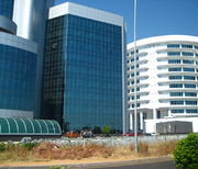 Two tall buildings. The right building is white and circular with about ten floors and with a grey trailer in front. The taller building on the left is cut off in the top right corner. It seems to be made up of smaller interconnected buildings with tinted windows