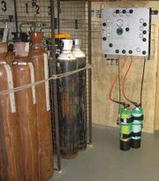 A panel on the wall is connected to diving cylinders by hoses. Nearby are several much larger cylinders, some painted brown and others black