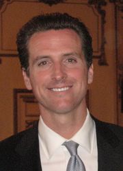 A white man wearing a suit and a tie stands in front of a wooden wall