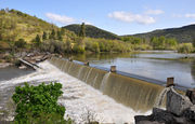 A large river flows over a dam and into a churning pool below. Green hills line the far shore.