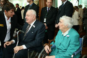 Whitlam, now in extreme old age, sits with an elderly lady as a woman bends to speak with him.  He holds a metal cane.  Other people, mostly men, stand behind him.