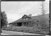 A black and white image of a long wooden building.  A single gable with two windows juts out from beneath a short chimney.  The porch is mostly enclosed with rough timbers.  The front of the building is lined with shrubbery and a stone wall.  Trees stand at either side of the structure.