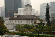 A two-storey neo-classical building showing Japanese architectural influences, with a central two-storey tower. In the foreground is a garden and tennis court and in the background are skyscrapers.