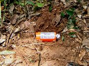 A red palm-sized cylindrical metal container, lying in a shallow freshly-dug hole in the ground, is labeled "ENREGISTREUR DE VOL / NE PAS OUVRIR".