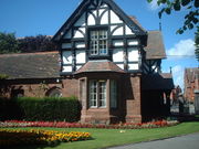 A two-storey house, the lower storey being in sandstone and the upper timber framed and gabled. To the right of the house are the entry gates to the park and in front of it are formal flower beds.