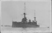 Photograph of a large warship. Along the bottom edge of the photograph, the phrases "HMAS AUSTRALIA" and "BUILT FOR THE AUSTRALIAN NAVY" are written in white. A sailboat and distant land can be seen in the left part of the photo.