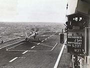 The deck of an aircraft carrier with a a propeller aircraft towards the front of the ship. Some of the ship's superstructure is at right, including a board displaying the wind conditions