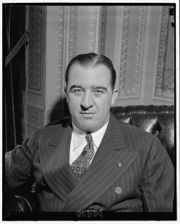 A man in his fifties, with thinning black hair, sitting at a desk, face-forward, wearing a suit