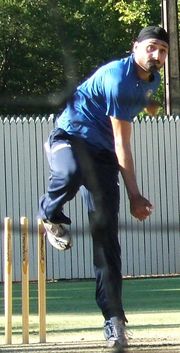 Man with black beard in a black turban, wearing navy tracksuit pants, blue t-shirt, follows through after bowling a ball in the cricket nets. His left leg is perpendicular to the ground, and his right leg is raised and bent back at the knee. His right bowling arm has rotated down to his side after releasing the ball.