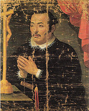 Painting  of an Asian man in black priest robes with white collar praying in    front of a crucifix.