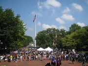 Red brick plaza, surrounded by trees with green leaves, with two white tents and an American flag flying from a flagpole in the center