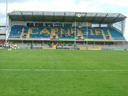 Inside a stadium with grass in the foreground and rugby posts and a large three-storey roofed stand with clock at the far end.  At ground-level there are steps and railings for standing patrons, and in front are a number of advertisements for the Leeds Building Society, PowerGen, Studio Jeff Banks, etc.  The upper part of the stand has about fifteen rows of seats, most of them coloured blue but the others collectively spelling out the word "Carnegie".  There are a small number of patrons in both sections and a few more on the top level in front of some windows at the rear.  To the left of the stand is part of another three-storey building.  In front of this, at the far left-hand end of the pitch, are a few drum-majorettes with pompoms.