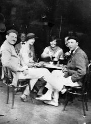 three men, dressed in light colored trousers and wearing hats, and two women, wearing light colored dresses, sitting at a sidewalk table