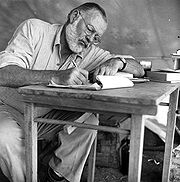 white-bearded man wearing light colored clothing writing at a small table