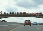 A bridge showcased against the sky, with the ground not visible. On the bridge, and lining it from left to right across the image are people, some holding Canadian flags.