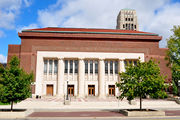 Red brick building with white stone facade. A tall white-colored stone clock tower with a green roof is in the background