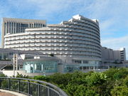 large circular multi-story building with stepped upper levels, facade consisting of multiple rows of balconies; foreground is a tree-lined park area, with a curved walkway railing, and additional buildings behind