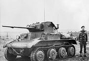 A small, squat tank sits in a field, its left-hand tracks facing the camera. Its gun barrel has been lengthened with a metal extension. A man in uniform with a black beret stands next to it. In the background, fields can be seen to the left, and several houses to the right.