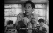 Photo of a government office in 1940's Tokyo: a bureaucrat, facing the camera and holding some kind of cloth in his right hand, stares beyond the left side of the frame as office workers appear behind him.