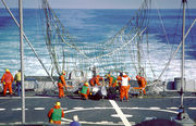 The rear deck of ship, with a large partially erect net visible near the center of the image. Many men in orange suits are working to free a white drone entangled in the net.