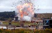 Viewed from a distance, with a telephoto lens, a large explosion is captured in its early stages.  In the foreground, assorted building materials are visible.  In the background, a hillside is partially covered by a forest.